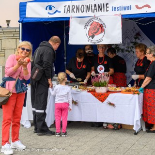 Festiwal Tarnowianie - dzień 2 - Na stoisku grupa kobiet w czarnych koszulkach, czerwonych wzorzystych spódnicach oraz z czerwonymi koralami na szyji, mężczyzna trzyma za rękę małą dziewczynkę i oglądają produkty, kobieta patrzy w obiektyw i uśmiechnięta trzyma w rękach dwa jabłka.  - Fot. Przemysław Sroka i Michał Żurowski