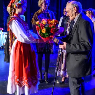 Festiwal Tarnowianie - dzień 3 - Dziewczyna w czerwono-białym stroju ludowym przekazuje bukiet kwiatów starszemu mężczyźnie w garniturze i okularach.  - Fot. Przemysław Sroka