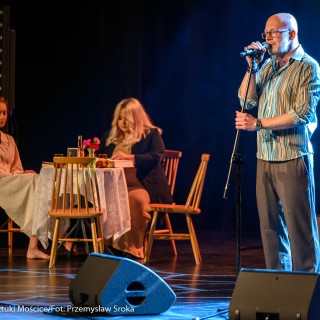 Festiwal Tarnowianie - dzień 3 - Mężczyzna w okularach i koszuli w paski stoi na scenie i śpiewa do mikrofonu, za nim w tle widać dwie kobiety siedzące przy stoliku.  - Fot. Przemysław Sroka