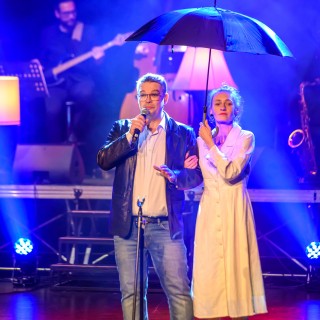 Festiwal Tarnowianie - dzień 3 - Na scenie kobieta w białej sukience stoi pod rękę z mężczyzną w okularach, który mówi do mikrofonu, w drugiej ręce trzyma parasol.  - Fot. Przemysław Sroka