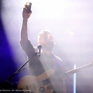 Koncert Kwiatu Jabłoni - Muzyk z gitarą i mikrofonem trzyma w wyciągniętej do góry dłoni telefon z zaświeconą latarką. - fot. Michał Żurowski
