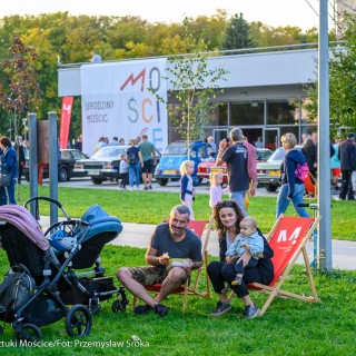 Urodziny Mościc - Grupa ludzi chodzi przed budynkiem CSM, mężczyzna oraz kobieta z dzieckiem na kolanach siedzą na leżakach i uśmiechają się do obiektywu do zdjęcia.  - Fot. Przemysław Sroka i Michał Żurowski