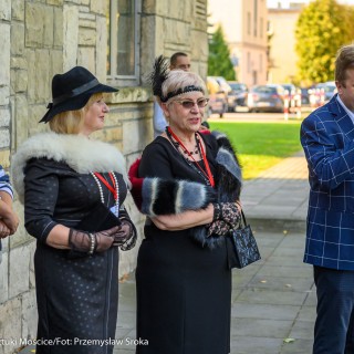 Urodziny Mościc - Trzy kobiety w eleganckich sukienkach oraz mężczyźni w garniturach stoją przed ścianą budynku CSM, jeden z nich trzyma w ręce mikrofon i mówi do niego.  - Fot. Przemysław Sroka i Michał Żurowski