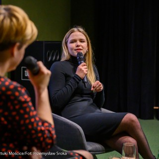 "Wszystkie nasze strachy" - spotkanie z Agatą Łabno - Na scenie siedzi Agata Łabno w czarnej sukience i mówi do mikrofonu. Z lewej strony widoczna jest od tyłu kobieta prowadząca spotkanie, która również trzyma mikrofon. - Fot. Przemysław Sroka