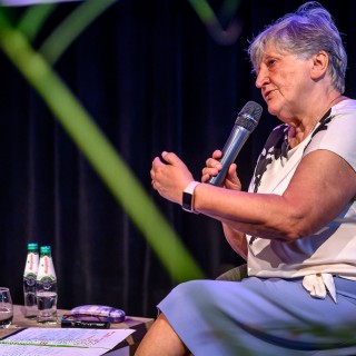Senior w Centrum - Narodowe Czytanie - "Nad Niemnem" - Anna Krakowska siedzi na scenie z mikrofonem w dłoni i mówi do słuchaczy. - Fot: Przemysław Sroka