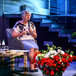 Senior w Centrum - Narodowe Czytanie - "Nad Niemnem" - Anna Krakowska siedzi na scenie z mikrofonem w dłoni i przemawia do słuchaczy. Na pierwszym planie widać wazon z białymi i czerwonymi kwiatami. - Fot: Przemysław Sroka