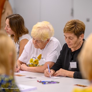 Warsztaty poetyckie Źródło - Uczestnicy warsztatów siedzą obok siebie przy stole. Dwie kobiety trzymają w dłoni długopis i piszą na kartce. - Fot: Przemysław Sroka