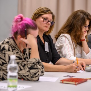 Warsztaty poetyckie Źródło - Trzy kobiety siedzą przy stole. Dwie z nich podpierają głowę ręką, jedna pisze na kartce.  - Fot: Przemysław Sroka