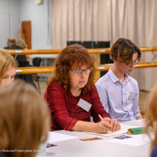 Warsztaty poetyckie Źródło - Uczestnicy warsztatów siedzą obok siebie przy stole. Jedna z kobiet trzyma długopis w dłoni i pisze na kartce. - Fot: Przemysław Sroka