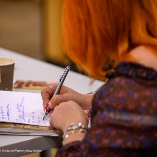 Warsztaty poetyckie Źródło - Kobieta trzyma w dłoni pióro i pisze na kartce. - Fot: Przemysław Sroka