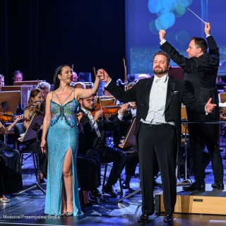 Koncert Sylwestrowy - Filharmonia Krakowska - Kobieta i mężczyzna stoją na scenie i trzymają się za ręce. Za nimi dyrygent dyryguje orkiestrą. - Fot: Przemysław Sroka