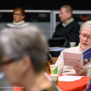 Senior w Centrum - Kolędowanie - Kobieta siedzi przy stole, trzyma śpiewnik w dłoni i śpiewa. - Fot: Przemysław Sroka