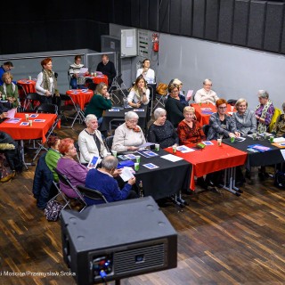 Senior w Centrum - Kolędowanie - Uczestnicy wydarzenia siedzą przy stołach. Przed nimi stoi kobieta z mikrofonem w dłoni. - Fot: Przemysław Sroka