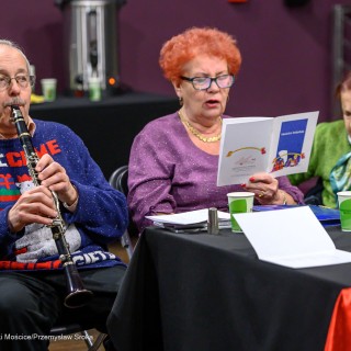Senior w Centrum - Kolędowanie - Dwie kobiety i mężczyzna siedzą przy stole. Jedna z kobiet trzyma w dłoniach śpiewnik. Mężczyzna gra na instrumencie. - Fot: Przemysław Sroka