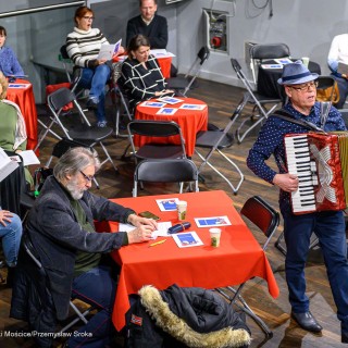 Senior w Centrum - Kolędowanie - Uczestnicy wydarzenia siedzą przy stołach i śpiewają. Między stołami stoi mężczyzna i gra na akordeonie. - Fot: Przemysław Sroka