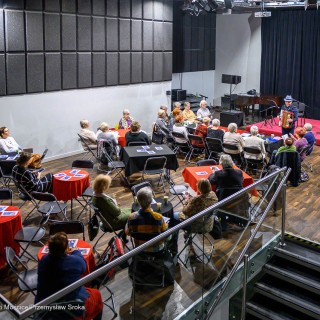 Senior w Centrum - Kolędowanie - Uczestnicy wydarzenia siedzą przy stołach. Przed nimi stoi mężczyzna z akordeonem, a na scenie stoją dwie dziewczynki w strojach ludowych. - Fot: Przemysław Sroka